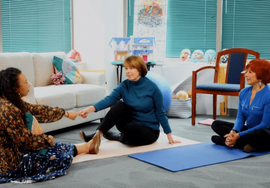 Woman holding hands across a yoga mat with therapist watching sitting on floor during therapy session