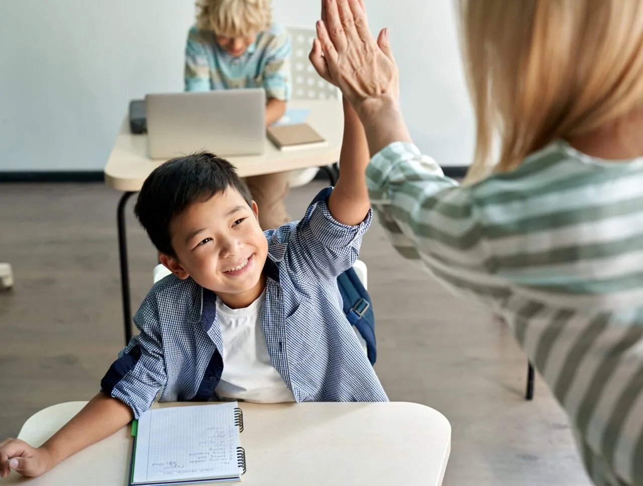 Teacher high fiving young Asian boy student sitting at desk smiling
