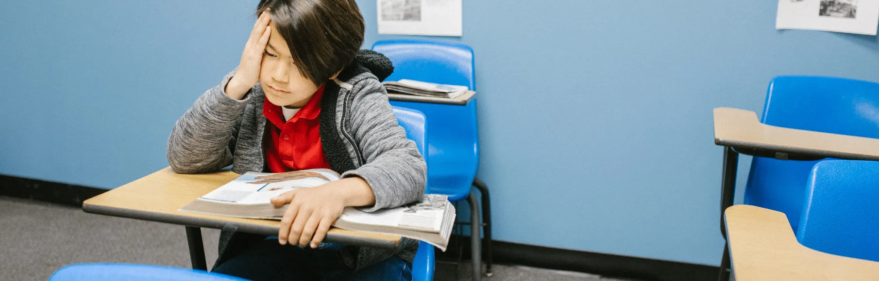 Asian boy looking sad sitting at desk with blue chair in school classroom