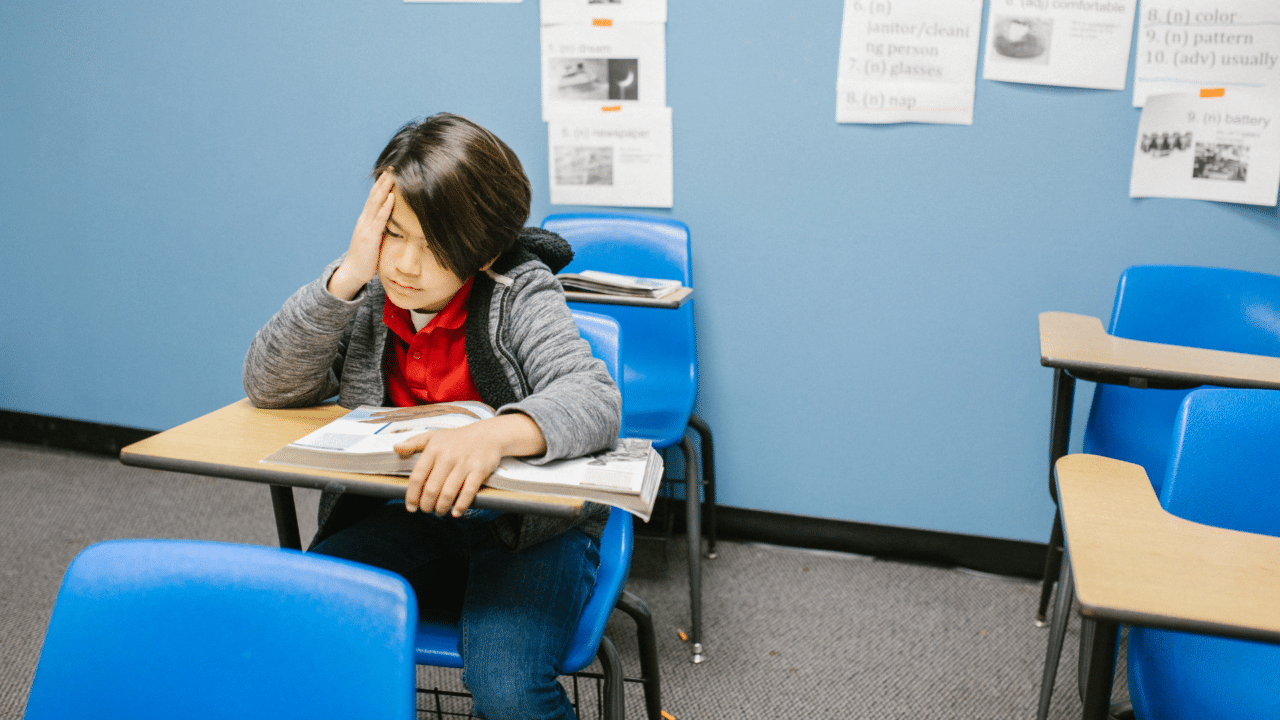 Asian boy looking sad sitting at desk with blue chair in school classroom