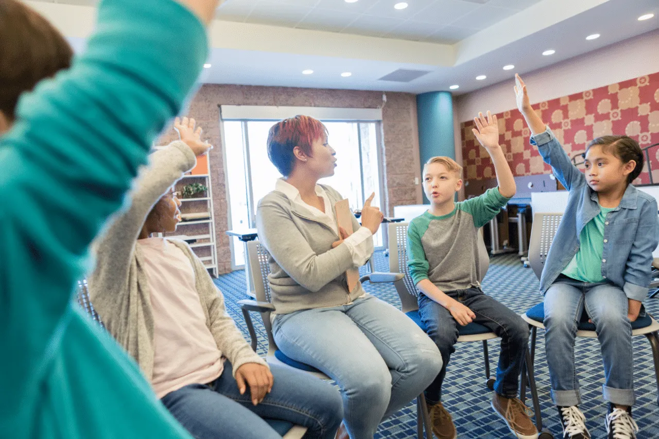 Multiracial woman sitting with diverse group of kids raising hands