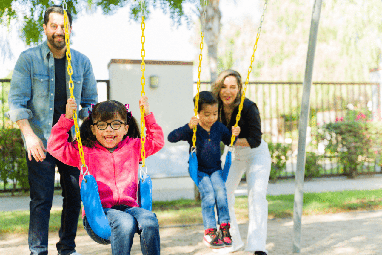 Two young children, boy and girl, swinging with parents pushing them on swing set