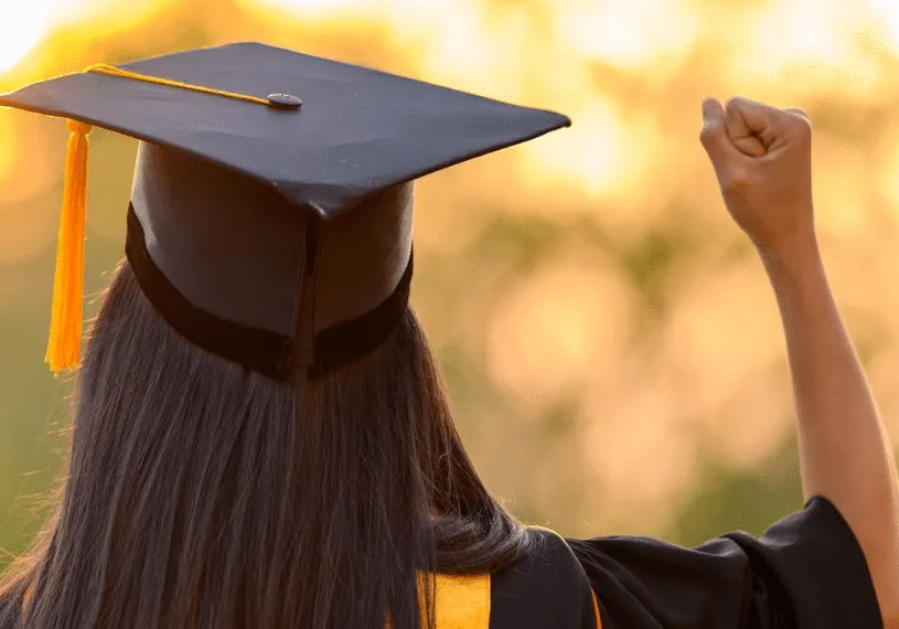 Girl facing away from camera wearing graduate cap and gown holding up right fist