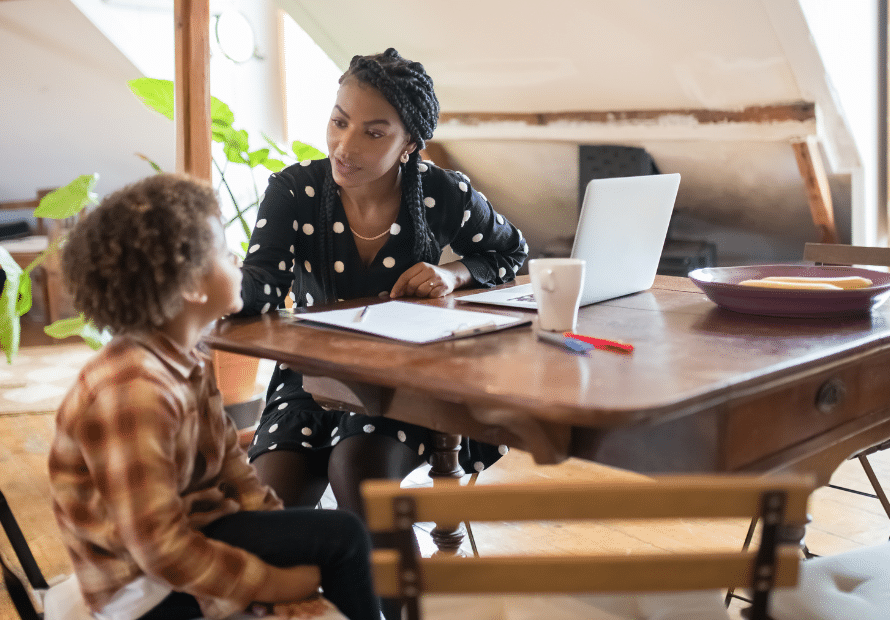 Black woman sitting at table with laptop and papers on desk with young biracial boy sitting next to her