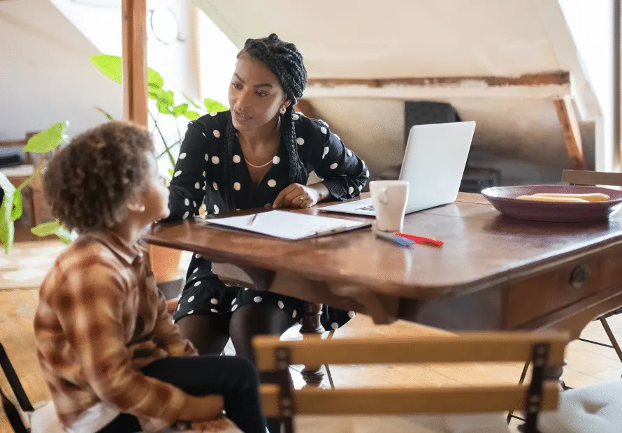 Black woman sitting at table with laptop and papers on desk with young biracial boy sitting next to her