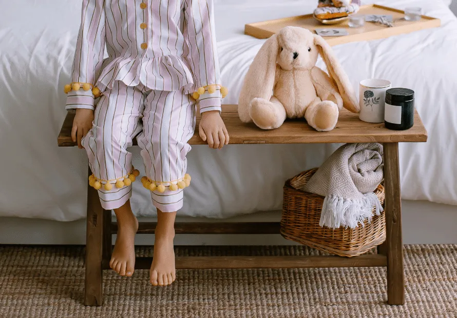 Young girl (no face showing) sitting on bench by bed with stuffed animal rabbit sitting next to her