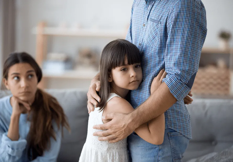 Young girl with sad face hugging Dad (no face shown). Mom in background looks tired and stressed.