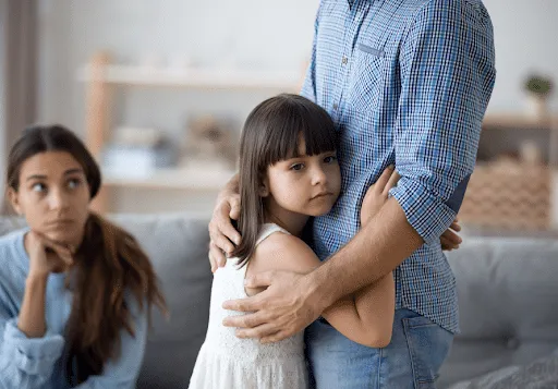 Young girl with sad face hugging Dad (no face shown). Mom in background looks tired and stressed.