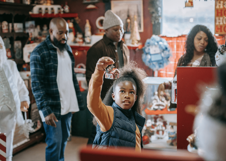 Young black girl holding up a house ornament in a store with other adults in the background