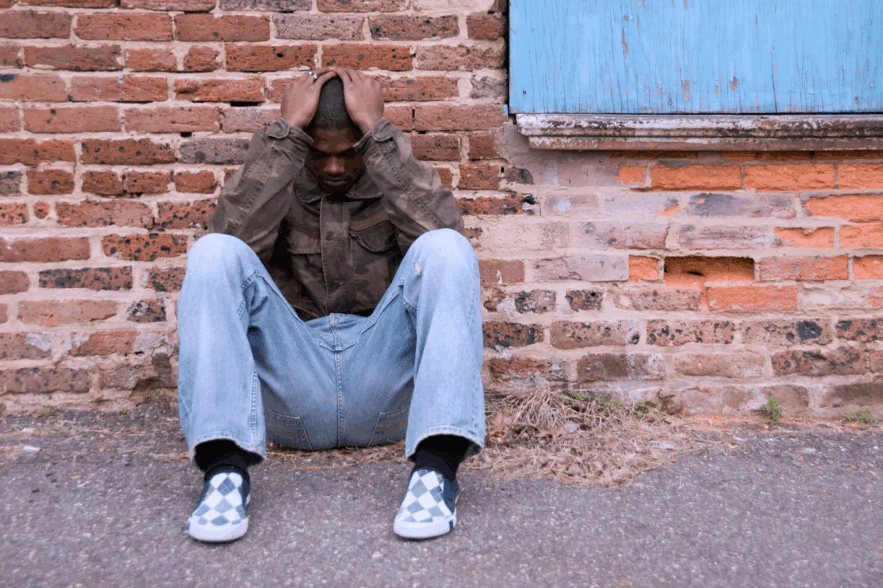 Young black male adult foster youth sitting on ground with hands covering forehead looking depressed